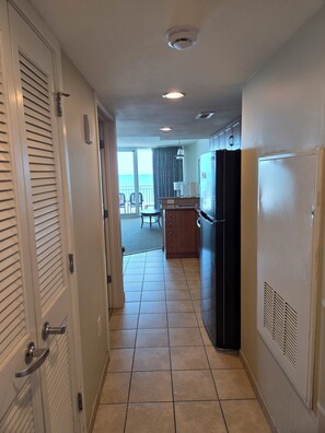 A corridor leading to a kitchen with a refrigerator, a dining area with a table and chairs, and a view of the ocean through a window.