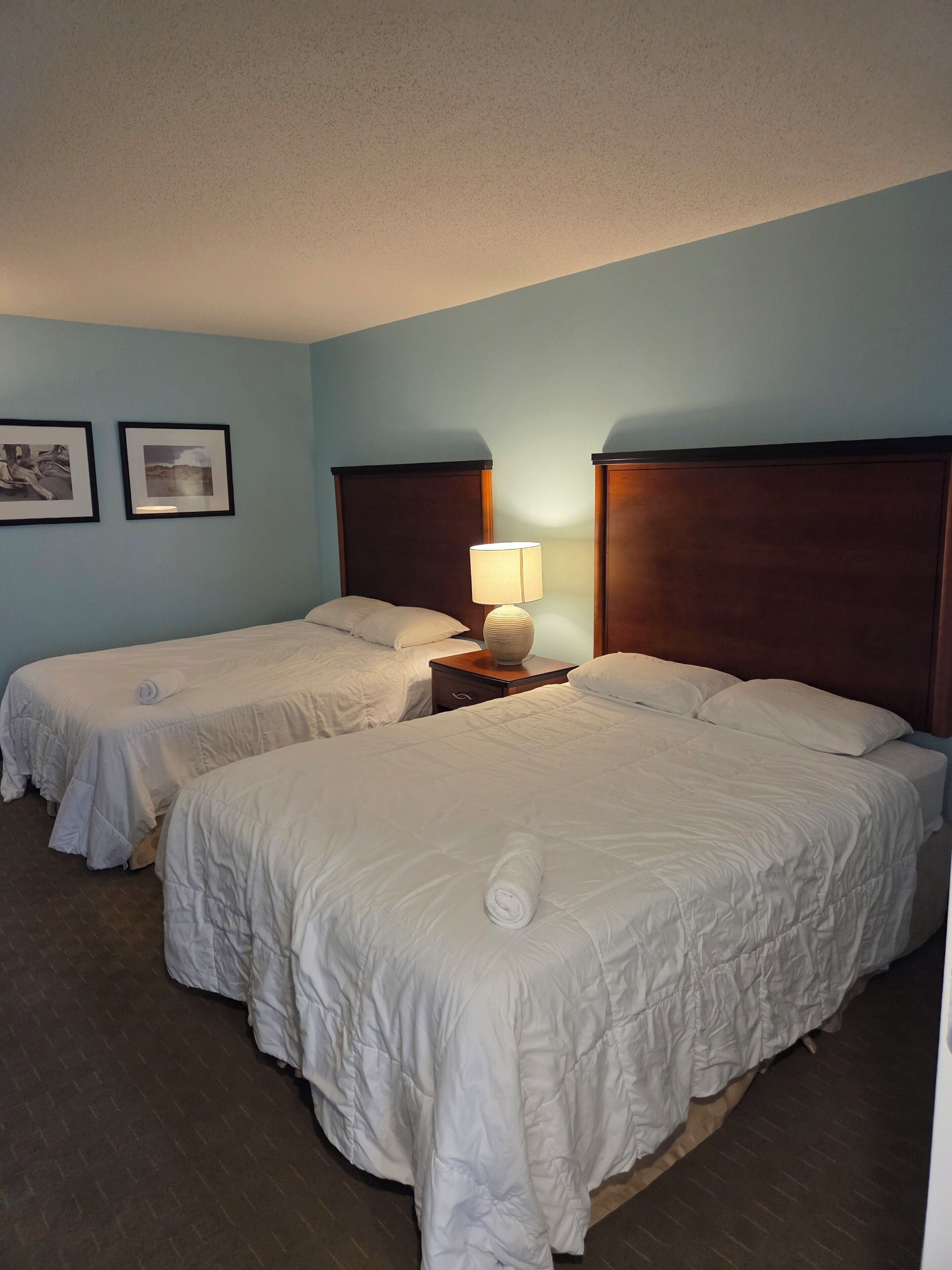 A hotel room with two beds, a nightstand with a lamp, and two framed pictures on the wall.