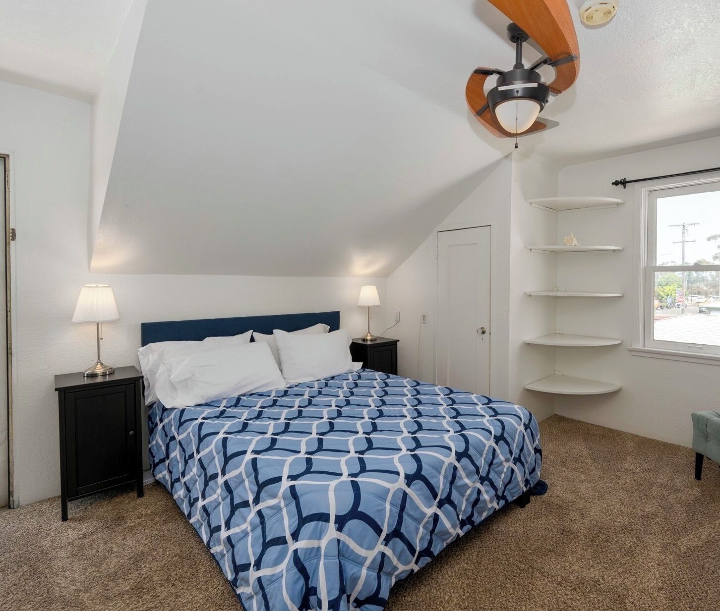 A bedroom with a bed, two bedside lamps, a ceiling fan, a window with a view, and a door.