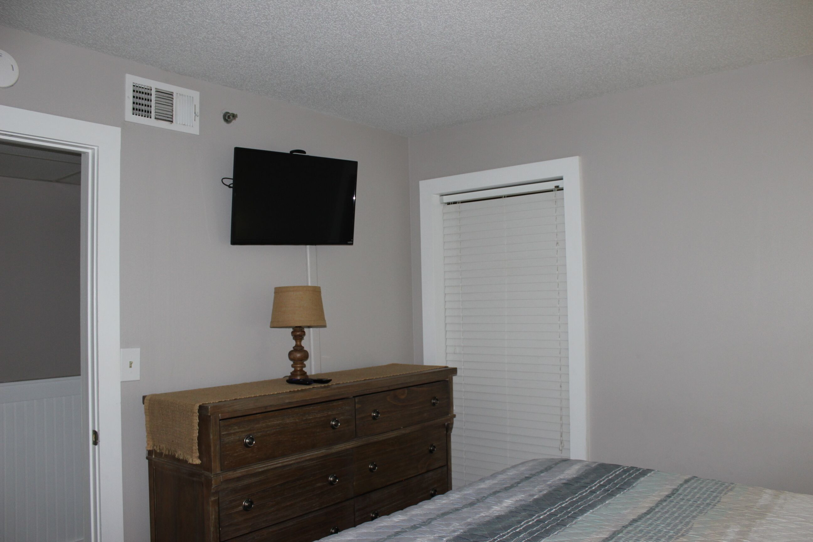 Une chambre à coucher comprenant un lit, une commode, un téléviseur fixé au mur et une fenêtre avec des stores.