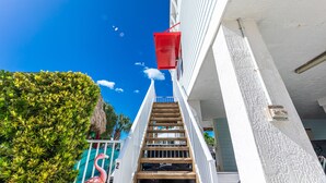 Eine Treppe führt zu einem Gebäude mit weißer Fassade, auf der Brüstung steht ein roter Eimer.