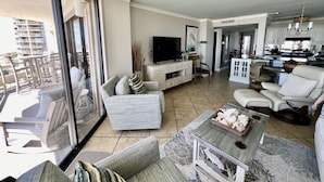 A spacious living room with a large TV, comfortable seating, a kitchen area, and a balcony with outdoor furniture.