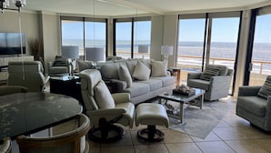 A spacious living room with a large window overlooking the beach, a comfortable sofa, a dining table, and a TV.
