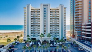 A multi-story hotel with a central entrance, parking lot, and a view of the beach.