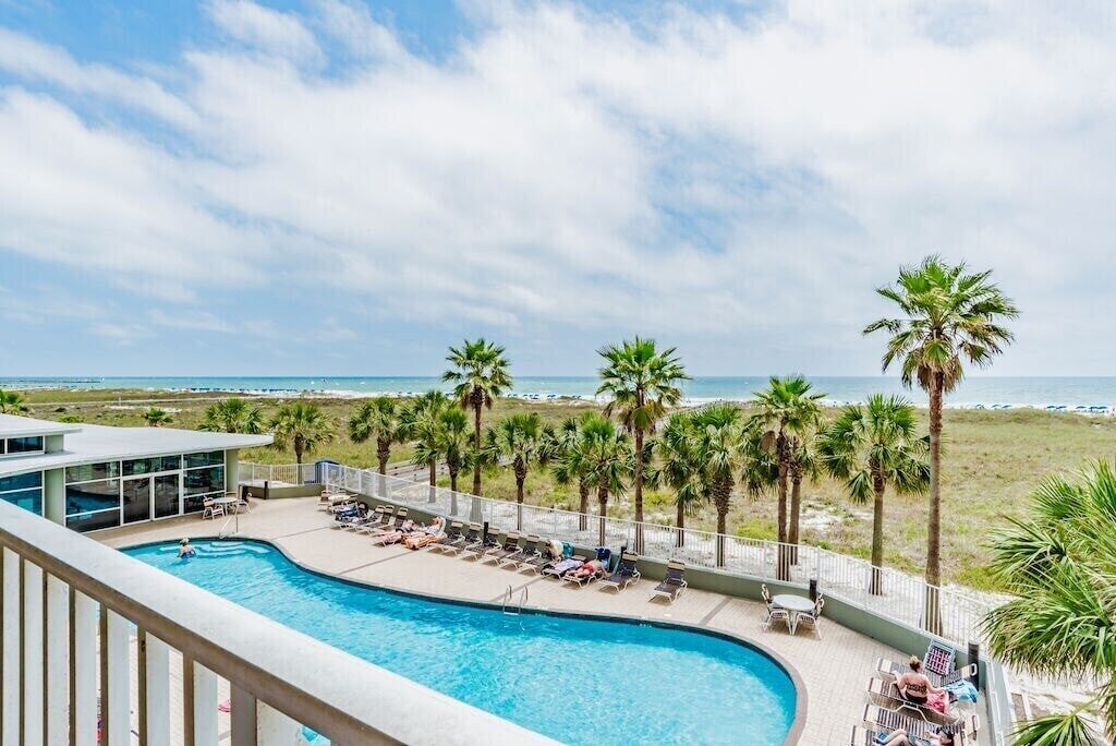 A pool area with lounge chairs and palm trees, overlooking a beach.