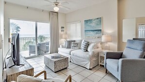 A spacious living room with a balcony, white furniture, a ceiling fan, and a large painting of the ocean.