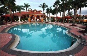 A circular pool with a surrounding deck, palm trees, and a building with a red roof in the background.