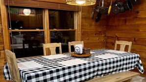 A wooden interior with a checkered tablecloth-covered table, chairs, a hanging light fixture, and a window with a view. - Log Cabin On 5 Private Acres, Gated Community With River Access (Rutherfordton)
