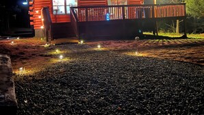 A wooden cabin with a porch and string lights, surrounded by gravel and trees. - Log Cabin On 5 Private Acres, Gated Community With River Access (Rutherfordton)