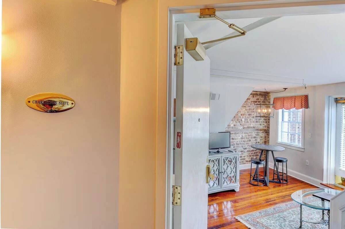 Une chambre d’hôtel avec une porte blanche, une pancarte indiquant qu’il s’agit de la « SUITE CHARLEMONT », un mur en briques, un téléviseur, une table à manger avec des chaises et un tapis à motifs.