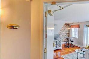 A hotel room with a white door, a sign indicating it's a 'CHARLEMONT SUITE,' a brick wall, a television, a dining table with chairs, and a patterned rug.