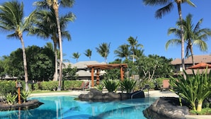 Piscina azul e límpida rodeada de pedras e palmeiras, com um pergolado de madeira e espreguiçadeiras ao fundo.