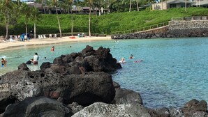 Uma praia com água cristalina, pessoas nadando e tomando sol, e um primeiro plano rochoso.