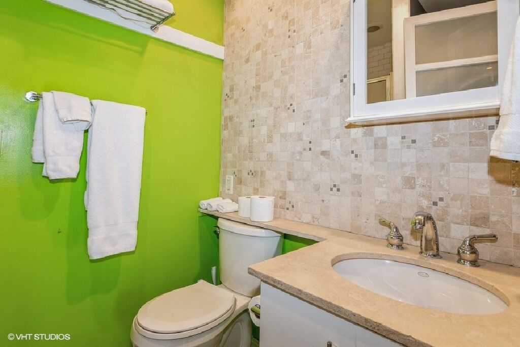 A bathroom with a green wall, a white toilet, a sink with a beige countertop, and a window above the sink.