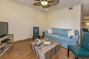 A living room with a television on a stand, a blue sofa, a wooden coffee table, and a ceiling fan.