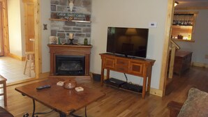 A cozy living room with a fireplace, a television on a wooden stand, and a stone wall with a sign reading "Cabin Sweet Cabin".