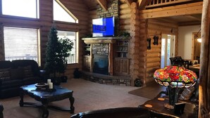 A log cabin interior with a stone fireplace, a television mounted above it, and a Christmas tree. - Luxury Log Home Retreat near Zion National Park (Toquerville)