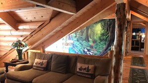 A cozy attic room with a wooden ceiling, a sofa with animal-themed cushions, a decorative lamp, and a large forest mural on the wall. - Luxury Log Home Retreat near Zion National Park (Toquerville)