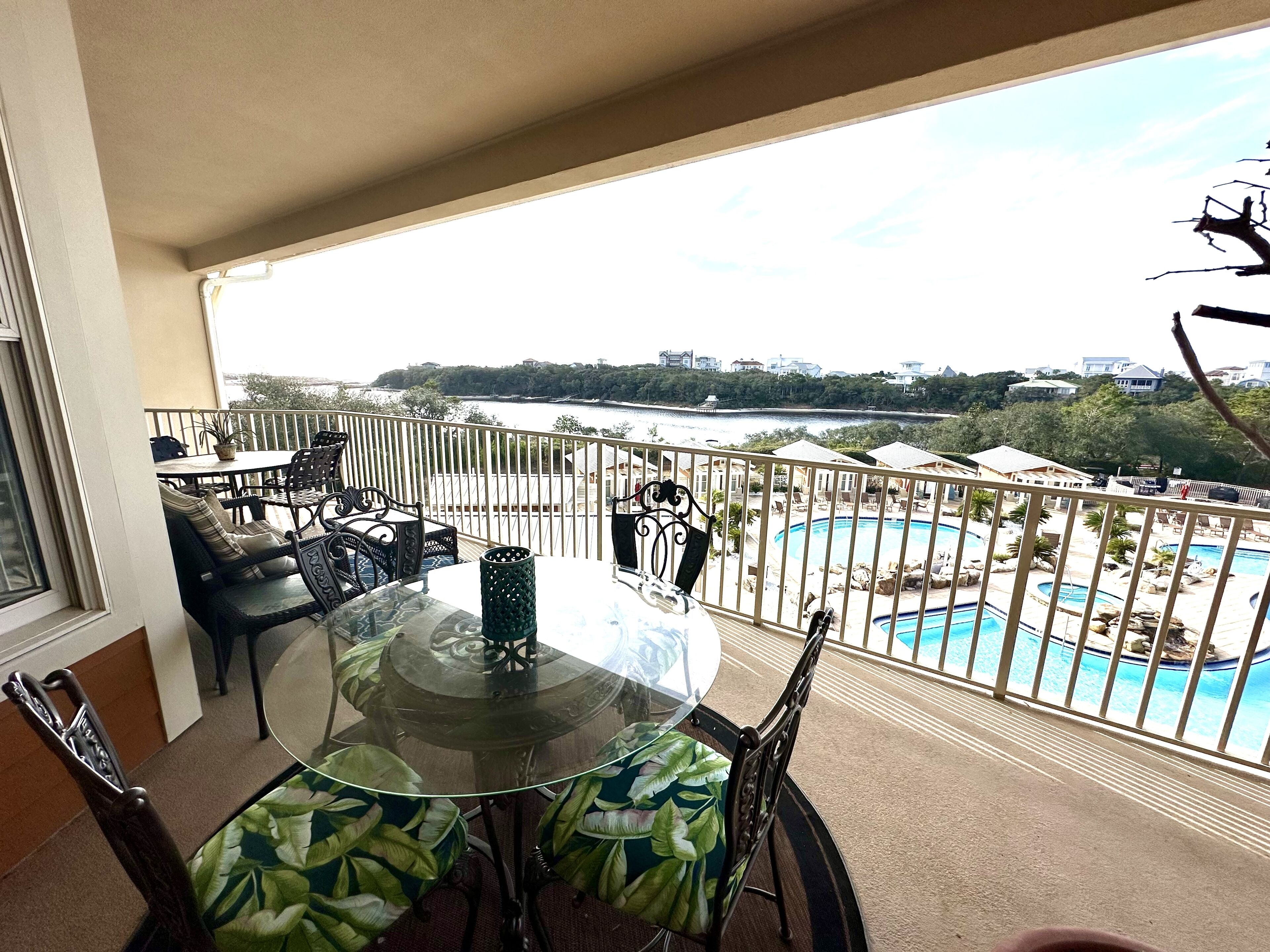 A balcony with a glass table and chairs, overlooking a swimming pool and a scenic view of a coastline.