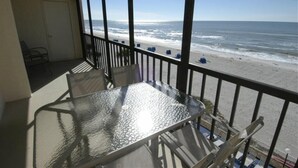 Un balcon avec une table et des chaises en verre, donnant sur une plage avec des vagues et un ciel dégagé.