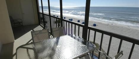 Un balcón con una mesa y sillas de vidrio, con vista a una playa y un cielo despejado.