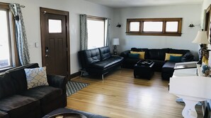 Sala de estar con un sofá, una mesita de centro y una mesita auxiliar blanca. Hay dos ventanas con cortinas, una puerta y una lámpara sobre la mesita.