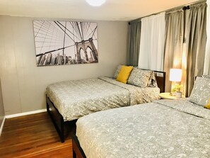 Quarto de hotel com duas camas, pintura de paisagem urbana na parede, abajur sobre a mesa de cabeceira e vista para o exterior através das cortinas.