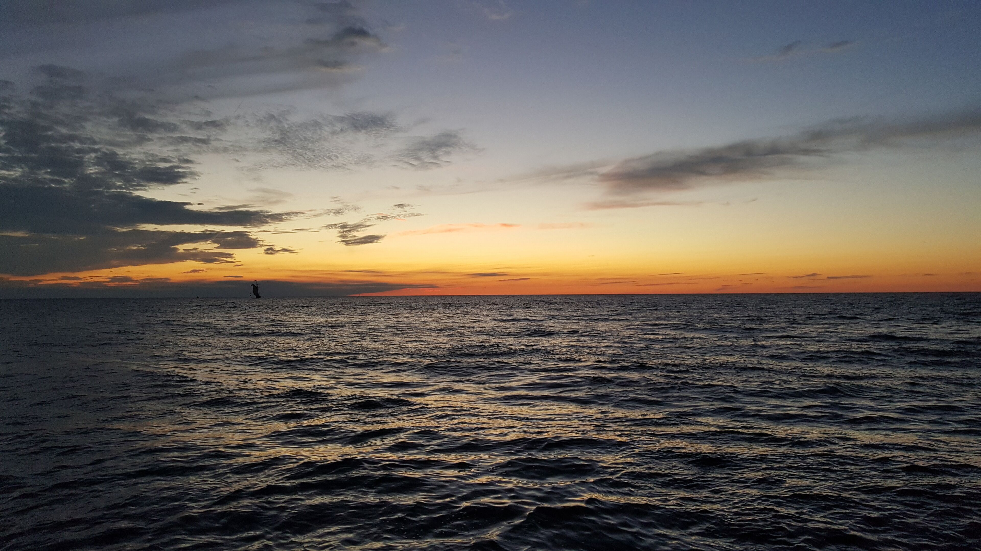 A view of the ocean at sunset with a distant sailboat.
