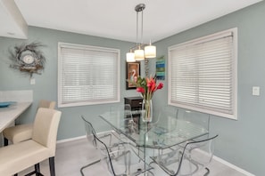 A dining area with a glass table and chairs, white blinds, and a vase with flowers.
