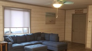 Una habitación con un sofá seccional, un ventilador de techo, una ventana con persianas y una puerta con un letrero.