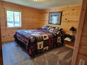 A cozy bedroom with a wooden log wall, a large bed with a colorful quilt, a nightstand with a lamp, and a window with a view of the outside. - Close to Wheeler's Point Boat Launch! (Baudette)