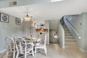 Un coin repas avec une table en verre et des chaises blanches, une cuisine à l’arrière-plan et un escalier.