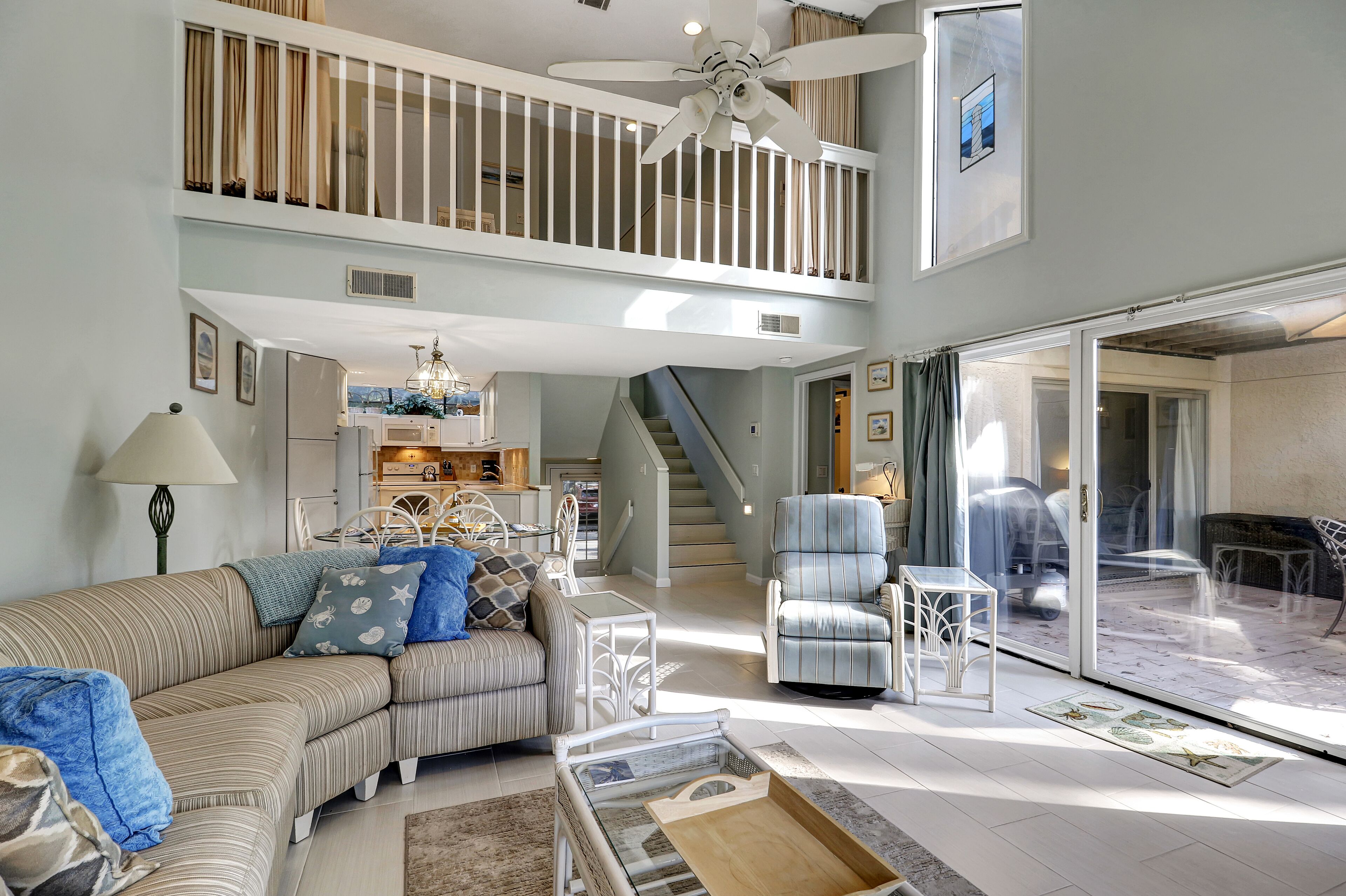 A spacious living room with a high ceiling, a ceiling fan, a staircase leading to an upper level, and a view of the outdoors through large sliding glass doors.