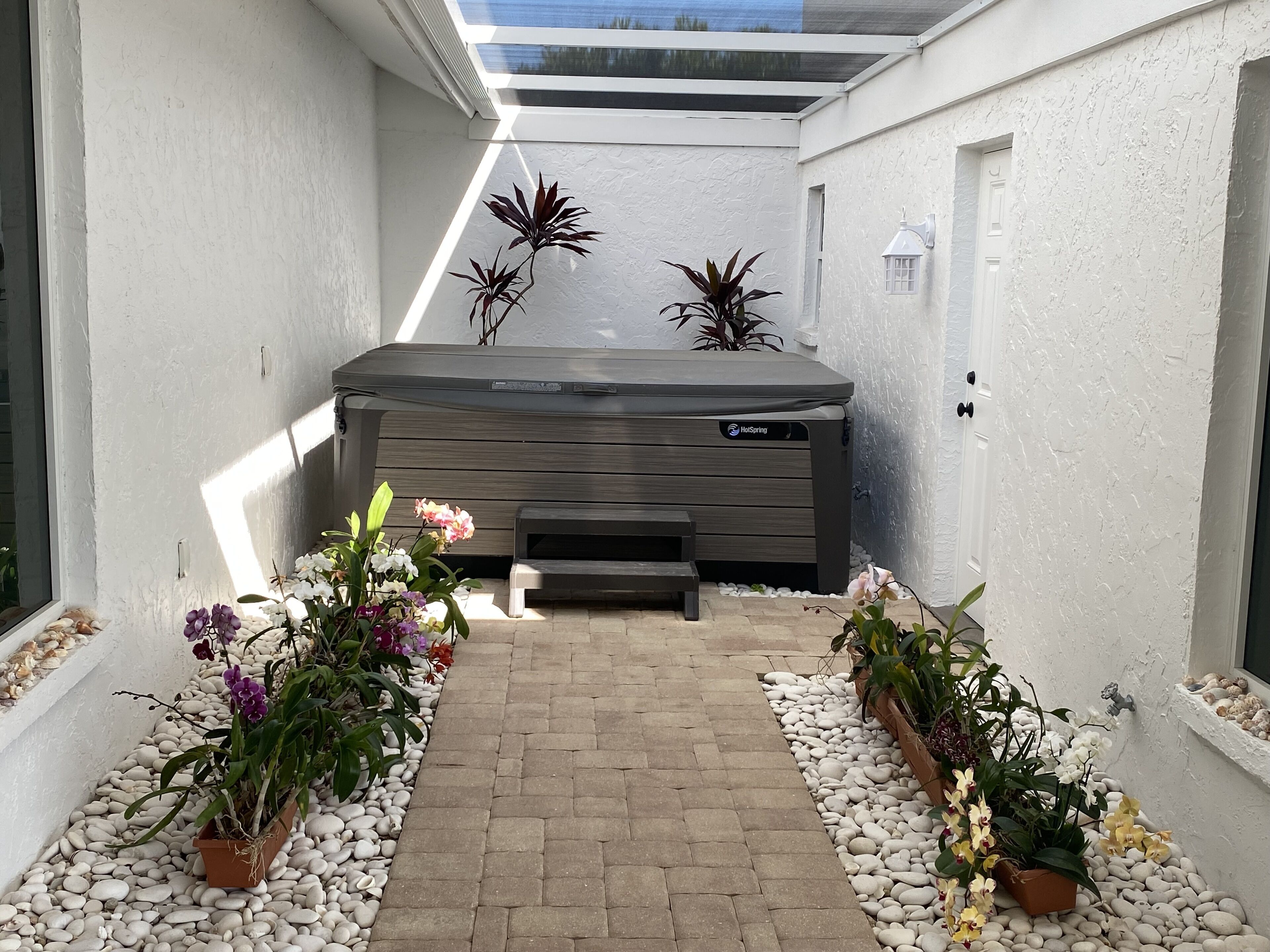 A narrow pathway leading to a hot tub with a wooden exterior, surrounded by white pebbles and potted plants with orchids.
