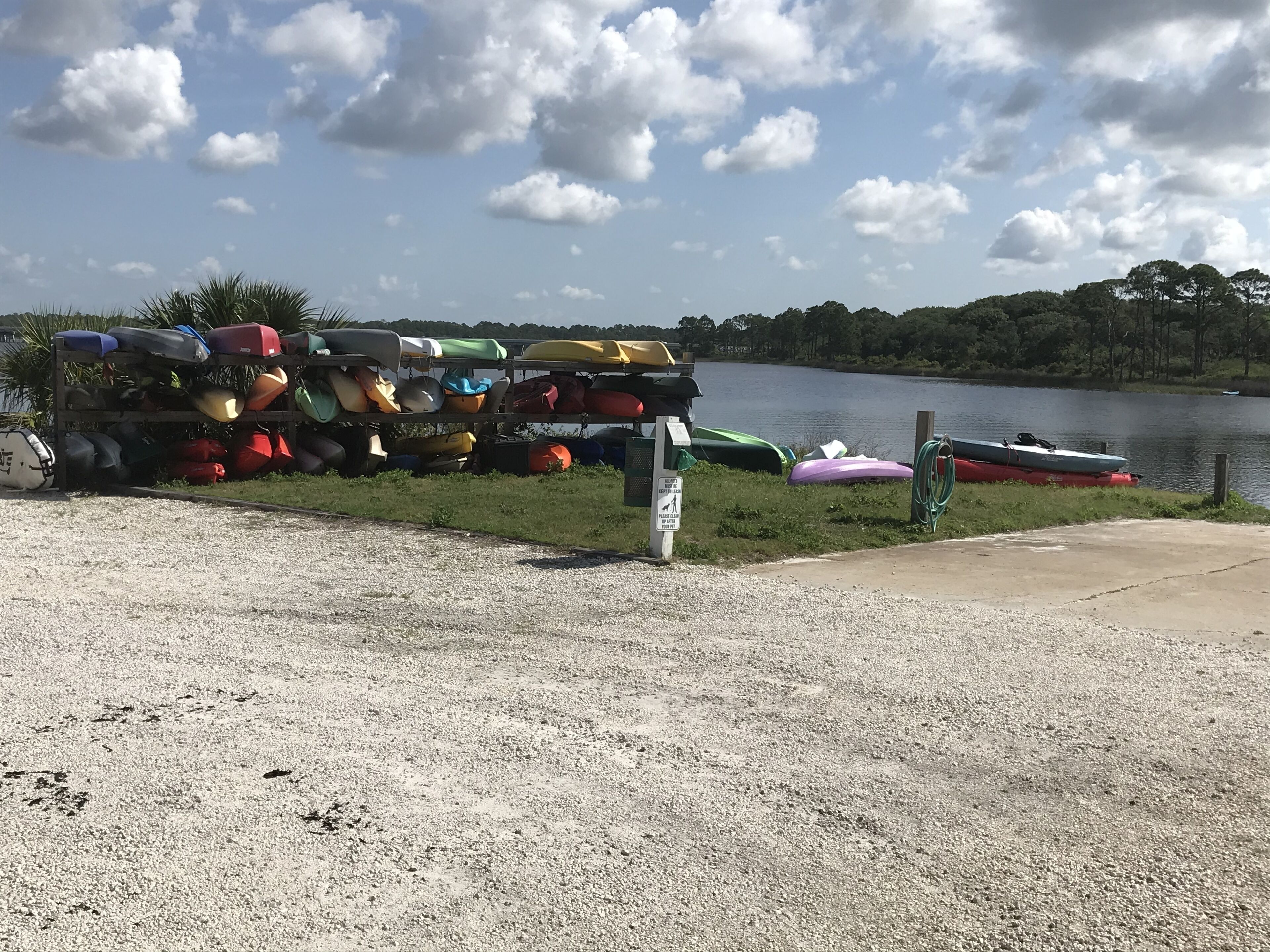 A gravel area with a rack of kayaks and canoes by a lake.
