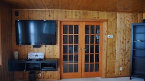Una habitación con paredes de paneles de madera, un televisor de pantalla plana grande montado en la pared, una puerta de madera con paneles de vidrio y un mueble oscuro debajo del televisor.