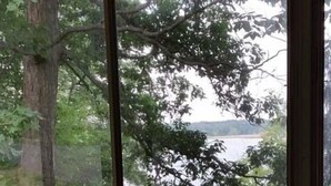 A room with a large window overlooking a lake, a guitar leaning against the window, and a chair in the foreground. - Secluded Lake Retreat-Exceptional Reviews! (Dowling)