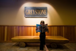 Uma pessoa lendo um livro em frente a um banco de madeira, com uma placa que diz "GREYSTONE" na parede ao fundo.