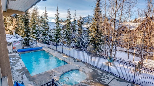 Piscina com hidromassagem, cercada por uma grade de metal, com paisagem nevada ao fundo, incluindo árvores e montanhas.