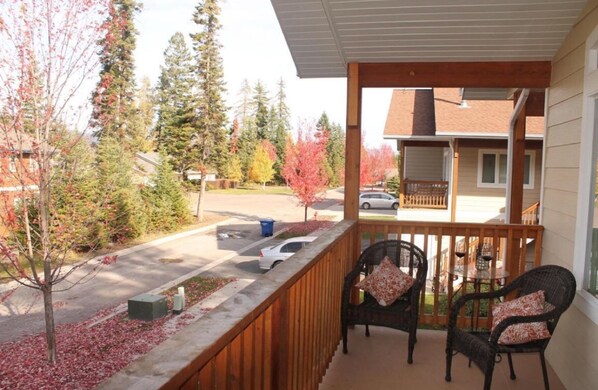 A wooden balcony with two wicker chairs and a small table, overlooking a street with autumn trees and a parked car.