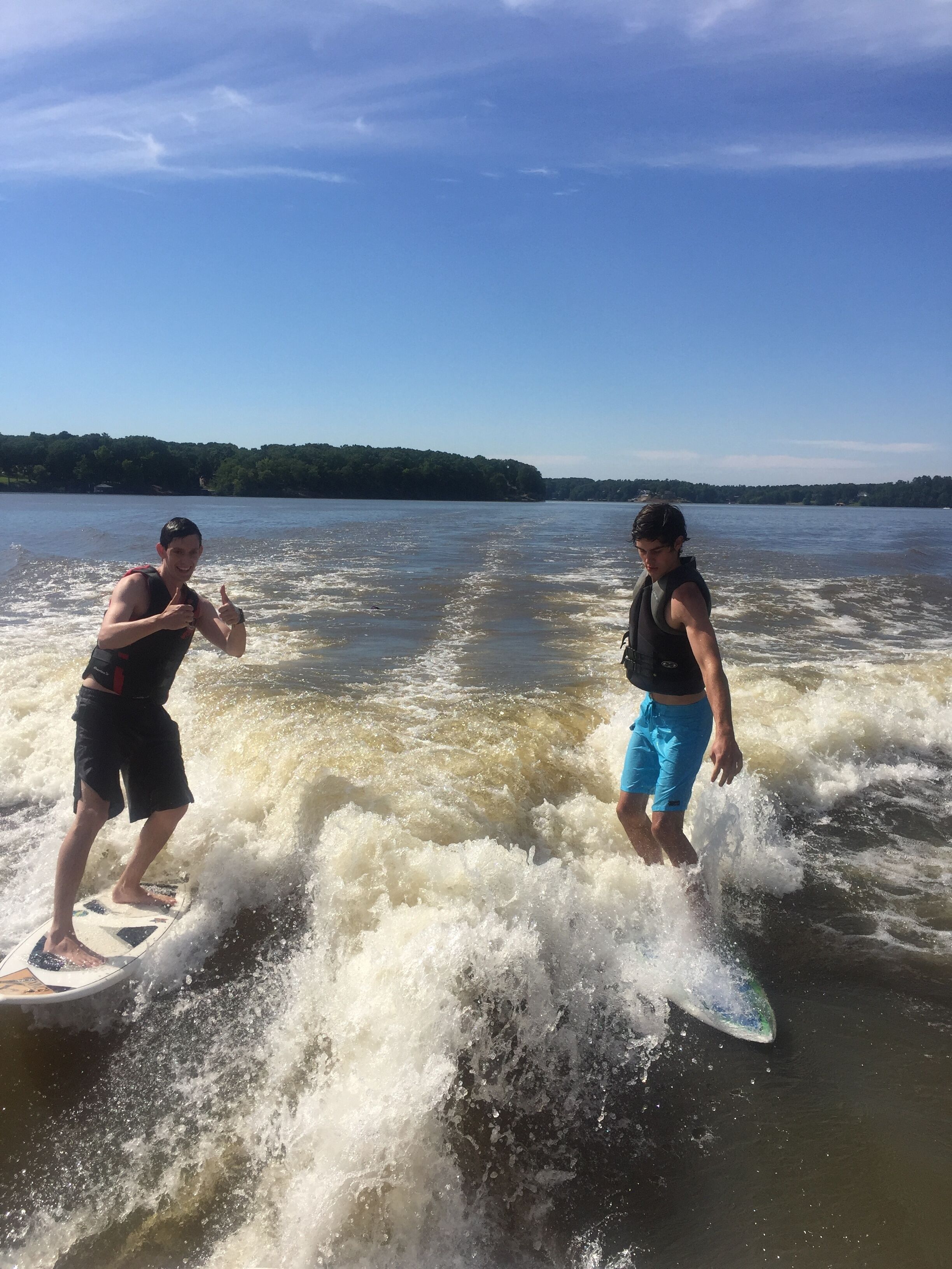 Two people wakeboarding on a river with a forested shoreline in the background.