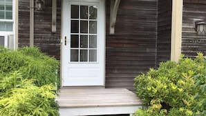 Ein Holzhaus mit weißer Tür, einer kleinen Veranda und einem Ziegelweg, der zum Eingang führt.