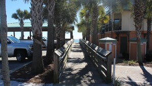 A wooden boardwalk with benches, palm trees, and a view of a building with balconies. - BEACH COTTAGE 3BR , GOLF CART W/HOUSE, PORCH SWING ,POOL 30 STEPS AWAY, PETS LOV (Destin)