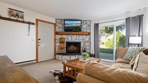 Ein gemütliches Wohnzimmer mit Kamin aus Stein, einem darüber montierten Fernseher und einem großen Fenster mit Blick ins Freie.