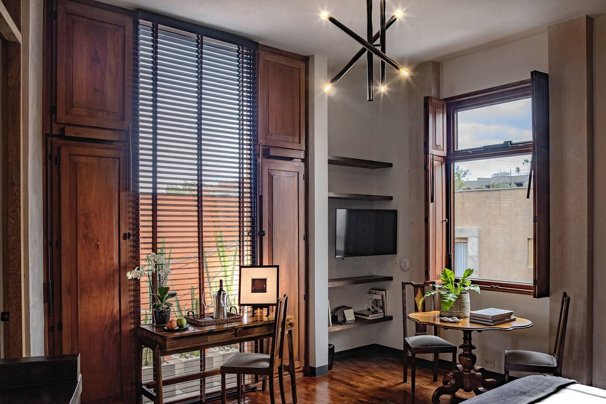 A hotel room with wooden flooring, a desk with a chair, a TV mounted on the wall, and a large window with a view of the outside.