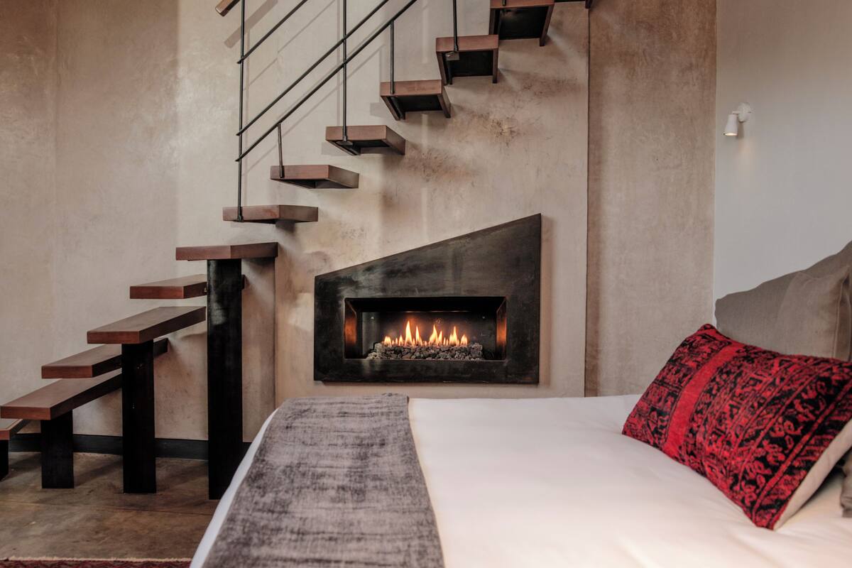 A modern bedroom with a fireplace, a staircase, and a bed with a red pillow.