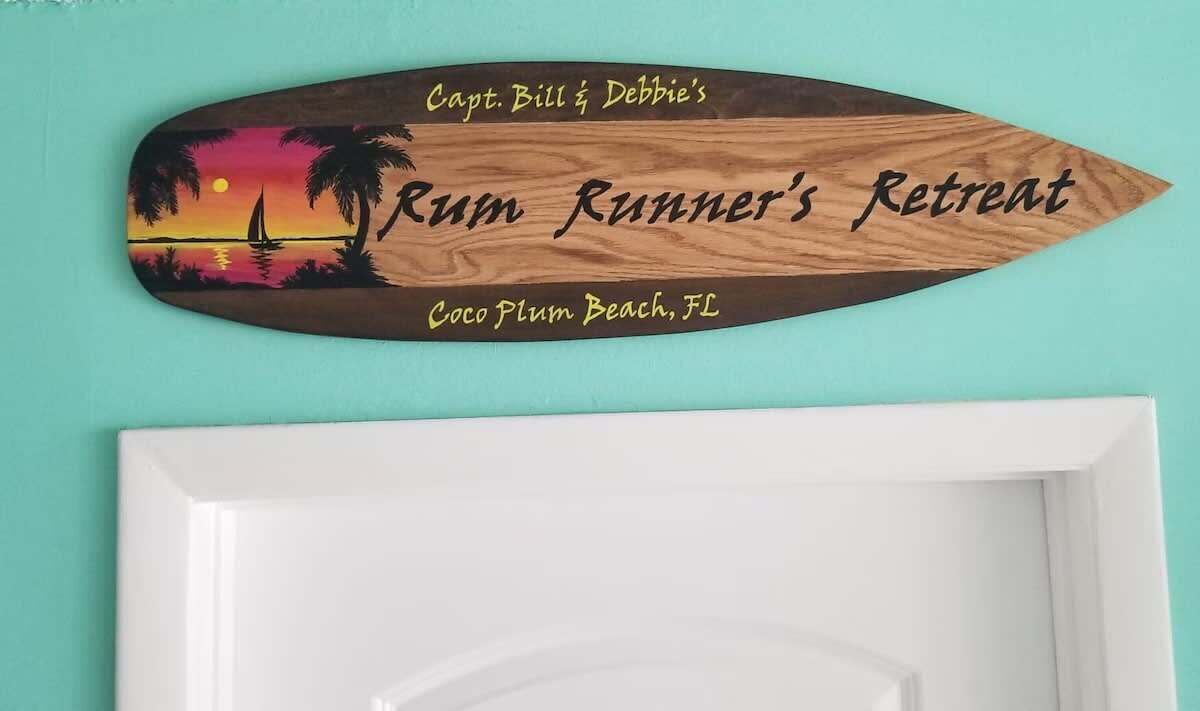 Puinen kyltti, jossa on kuva auringonlaskusta ja teksti, joka kertoo, että tässä on kapteeni Billin ja Debbie-vaimon ”Rum Runner’s Retreat” -paikka, Coco Plum Beach, Florida.
