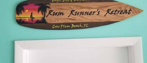 Puinen kyltti, jossa on kuva auringonlaskusta ja teksti, joka kertoo, että tässä on kapteeni Billin ja Debbie-vaimon ”Rum Runner’s Retreat” -paikka, Coco Plum Beach, Florida.