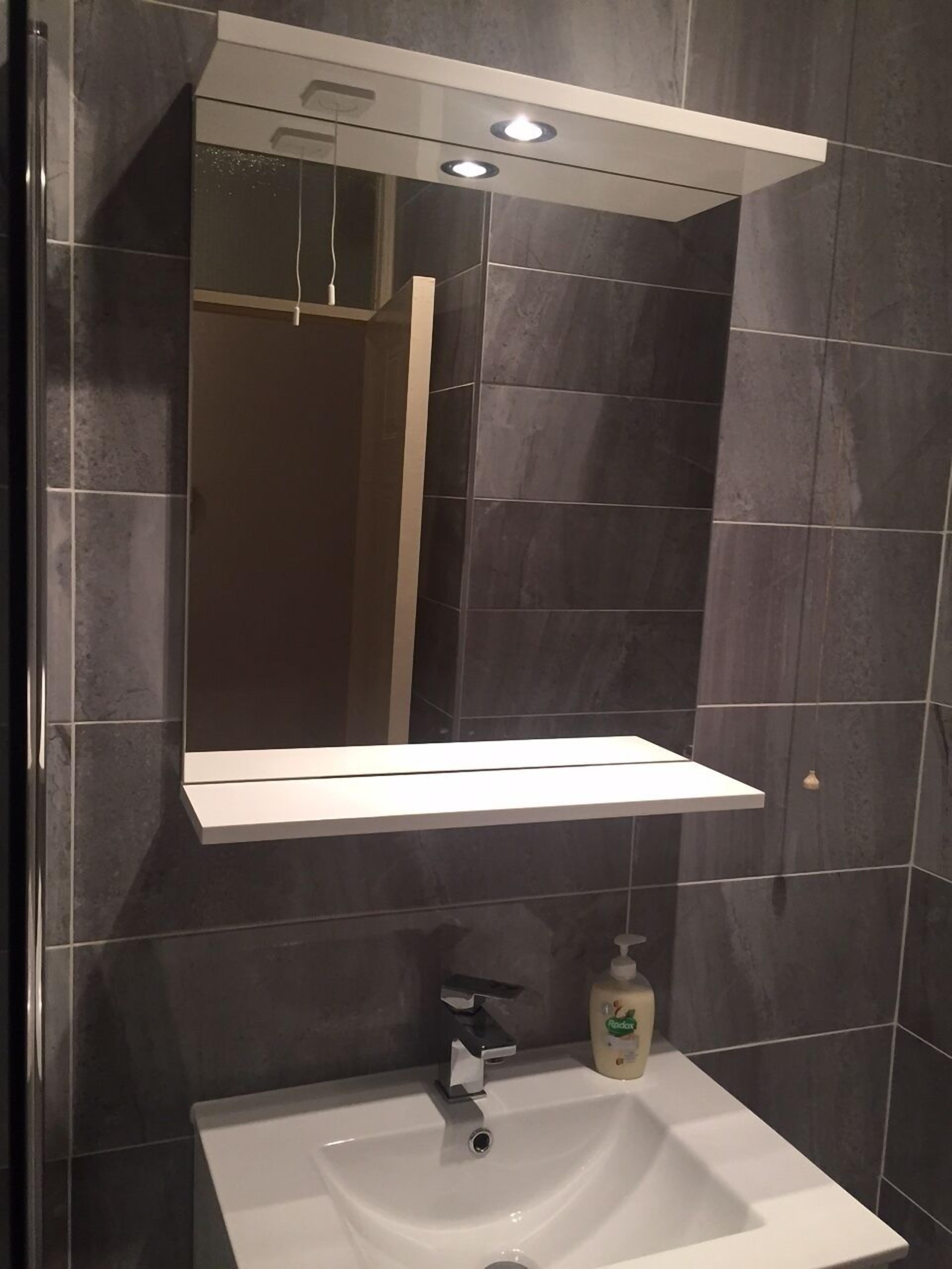 A bathroom with dark tiled walls, a white sink, a mirror above the sink, and a shelf above the mirror.