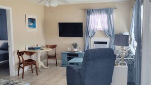A living room with a ceiling fan, a TV, a dining table with chairs, a blue cabinet, and a window with curtains. - Steps to Beach & Tranquility, #3 Walk to Spectacular Sunsets and Dining (Indian Rocks Beach)